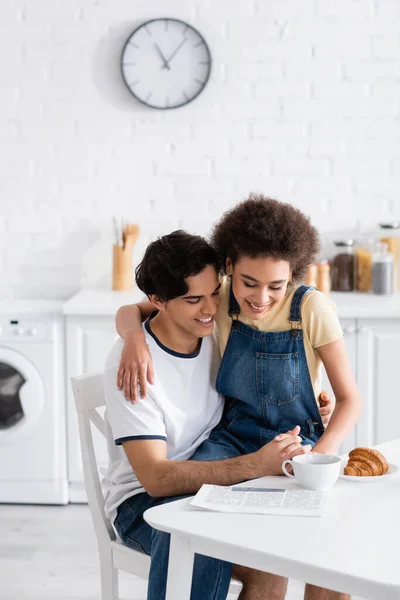 Feliz casal multiétnico de mãos dadas enquanto lê jornal durante o café da manhã — Fotografia de Stock