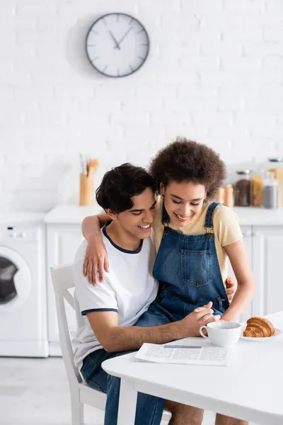 Feliz pareja multiétnica cogida de la mano mientras lee el periódico durante el desayuno - foto de stock