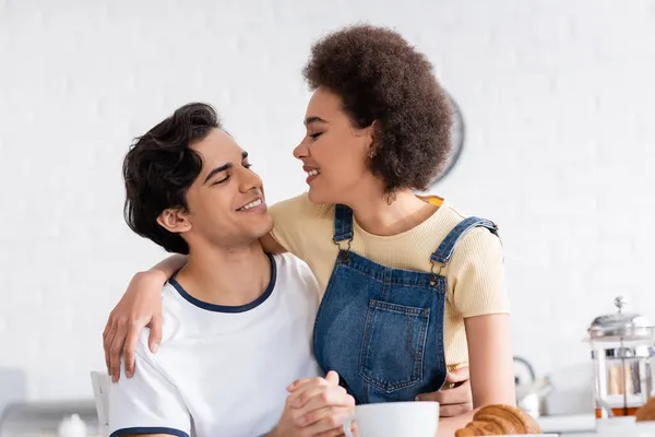 Feliz pareja multiétnica tomados de la mano mientras se miran durante el desayuno - foto de stock