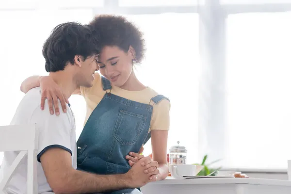 Feliz pareja multiétnica abrazándose y tomándose de la mano durante el desayuno - foto de stock