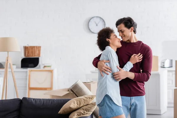 Cheerful multiethnic couple hugging and smiling during relocation to new home — Stock Photo