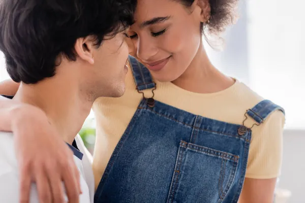 Feliz casal multiétnico abraçando e sorrindo em casa — Fotografia de Stock