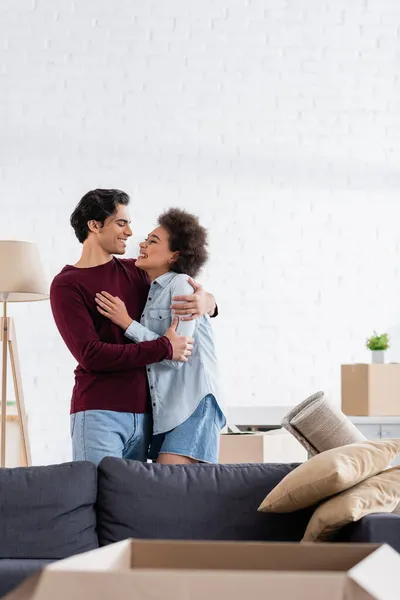 Feliz pareja multiétnica abrazando y sonriendo durante la reubicación a un nuevo hogar - foto de stock