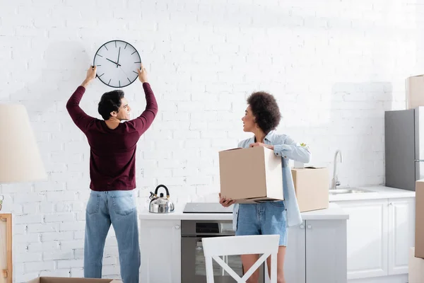 Homme heureux pendu horloge murale près de petite amie afro-américaine avec boîte en carton — Photo de stock