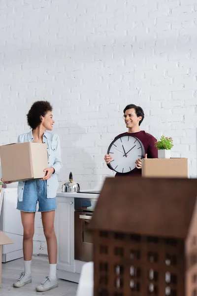 Glücklicher Mann hält Wanduhr nahe afrikanisch-amerikanischer Freundin mit Karton — Stockfoto