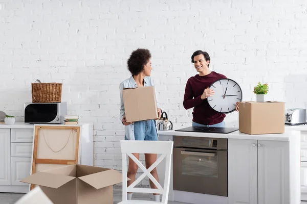 Fröhlicher Mann hält Wanduhr nahe afrikanisch-amerikanischer Freundin mit Karton — Stockfoto