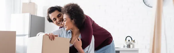 Feliz casal interracial abraçando perto caixa de papelão, banner — Fotografia de Stock