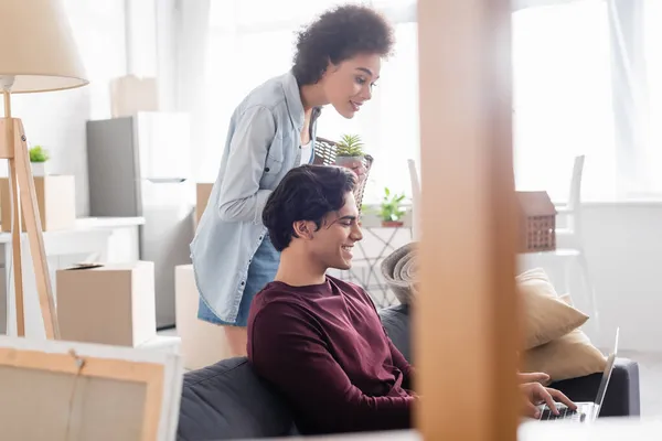 Cheerful african american woman holding basket near smiling boyfriend using laptop — Stock Photo