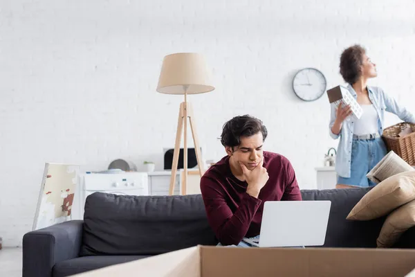 Freelancer using laptop near blurred african american woman holding basket and house model during relocation — Stock Photo
