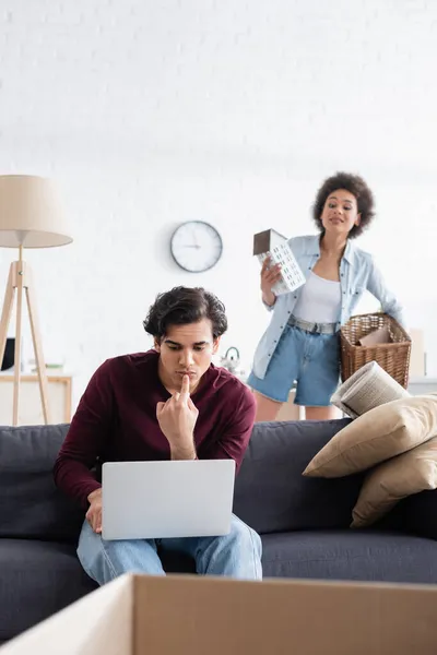 Pensive freelancer using laptop near curious african american woman holding basket and house model during relocation — Stock Photo