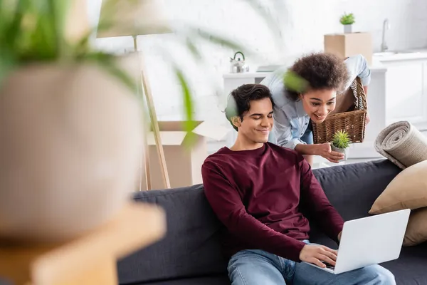 Allegra donna afro-americana che tiene cesto e pianta vicino al fidanzato sorridente utilizzando il computer portatile — Foto stock