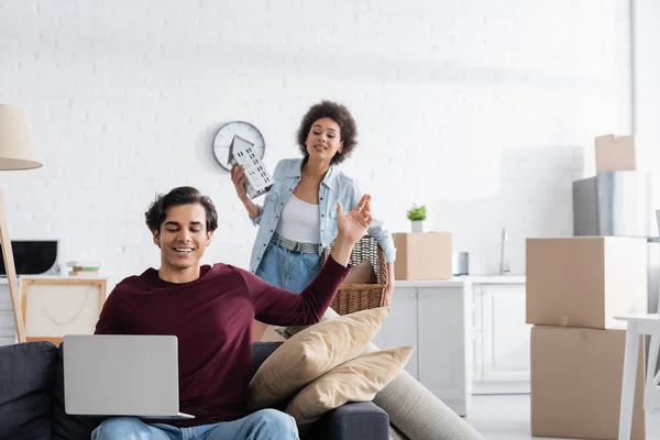 Fröhliche Freiberuflerin mit Laptop neben neugieriger Afroamerikanerin mit Korb und Hausmodell beim Umzug — Stockfoto