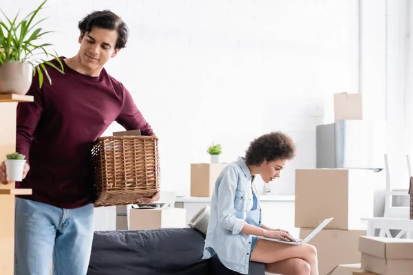 African american woman using laptop while man holding plant and basket — Stock Photo