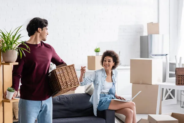 Mujer afroamericana feliz usando el ordenador portátil mientras gesticulaba cerca de hombre sosteniendo planta y cesta - foto de stock