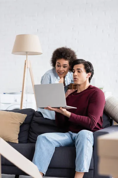 Mann zeigt fröhlicher Afroamerikanerin Laptop im Wohnzimmer — Stockfoto