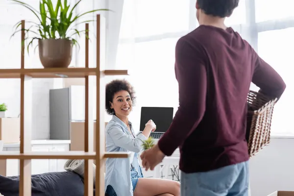 Glückliche afrikanisch-amerikanische Frau zeigt auf Laptop, während sie einen verschwommenen Mann mit Pflanze und Korb betrachtet — Stockfoto