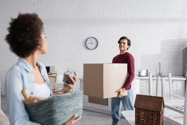 Happy man holding box near blurred african american girlfriend with basket — Stock Photo