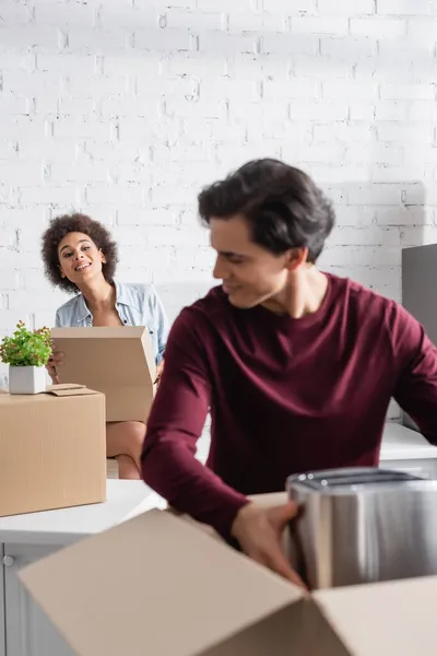 Borrosa joven hombre desembalaje caja con tostadora cerca curiosa afro-americana novia - foto de stock