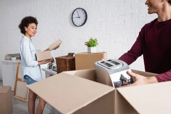 Happy young man unpacking box with toaster near smiling african american girlfriend — Stock Photo