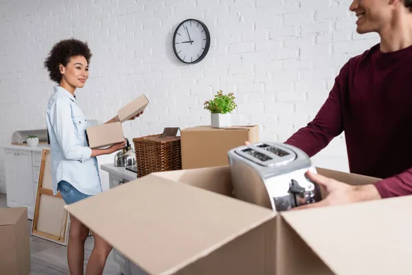 Feliz joven hombre desembalaje caja con tostadora cerca sonriente afro-americana novia - foto de stock