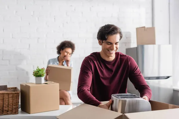 Cheerful young man unpacking box with toaster near blurred african american girlfriend — Stock Photo