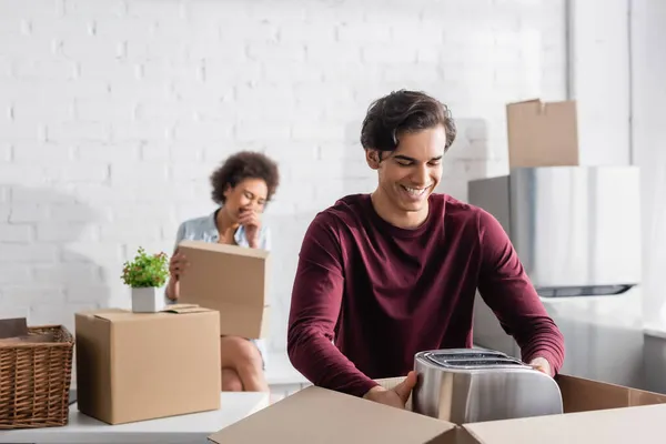 Alegre joven hombre desembalaje caja con tostadora cerca borrosa africana americana novia - foto de stock