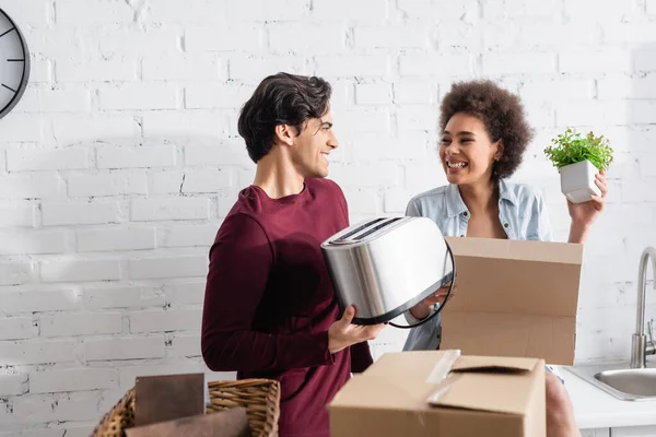 Happy young man holding toaster near smiling african american girlfriend with plant — Stock Photo