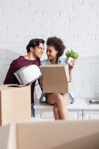 Happy young man holding toaster near joyful african american girlfriend with plant — Stock Photo