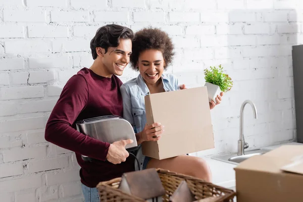 Cheerful young man holding toaster near joyful african american girlfriend with plant — Stock Photo