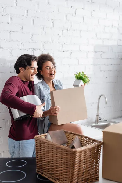 Cheerful young man holding toaster near happy african american girlfriend with plant in kitchen — Stock Photo