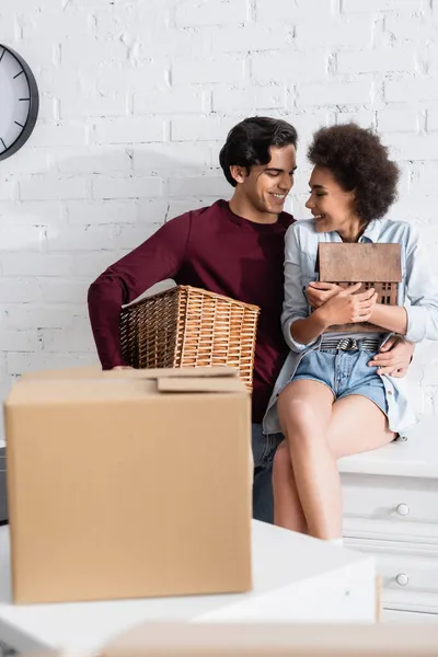 Fröhlicher junger Mann hält Korb in der Nähe glücklicher afrikanisch-amerikanischer Freundin mit Hausmodell — Stockfoto