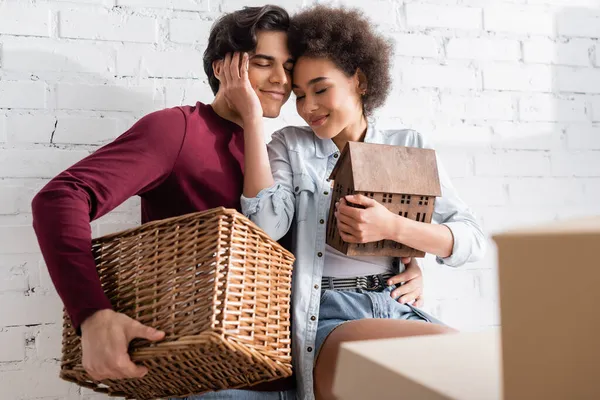 Joyeux jeune homme tenant panier près heureux copine afro-américaine avec modèle de maison — Photo de stock