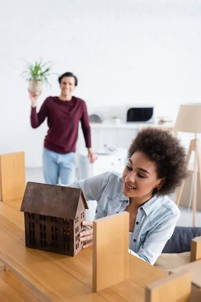 Feliz africano americano mujer colocación de madera casa modelo en rack cerca borrosa novio en fondo - foto de stock