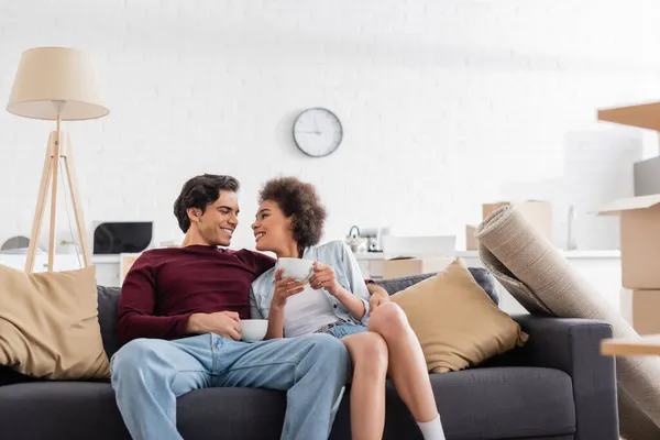 Feliz casal multiétnico segurando xícaras de chá enquanto descansa no sofá durante a realocação — Fotografia de Stock