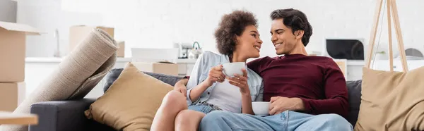 Feliz casal multiétnico segurando xícaras de chá enquanto descansa no sofá durante a realocação, banner — Fotografia de Stock
