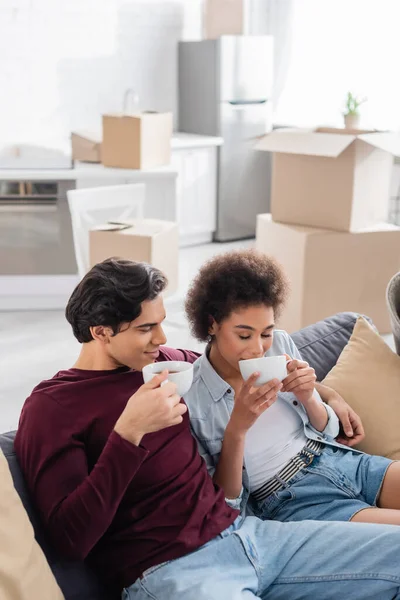 Happy multiethnic couple drinking tea while resting on couch during relocation — Stock Photo