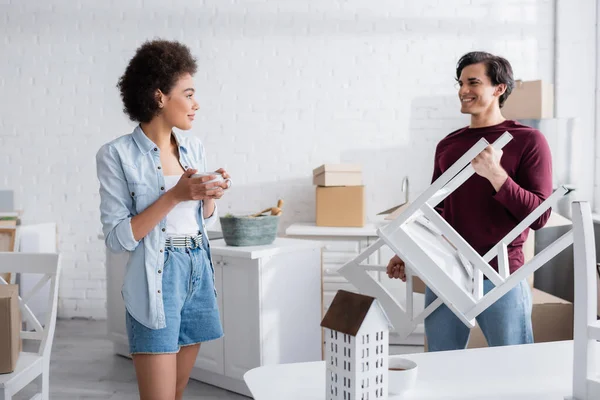 Happy african american woman holding cup of tea and looking at boyfriend with chair — Stock Photo