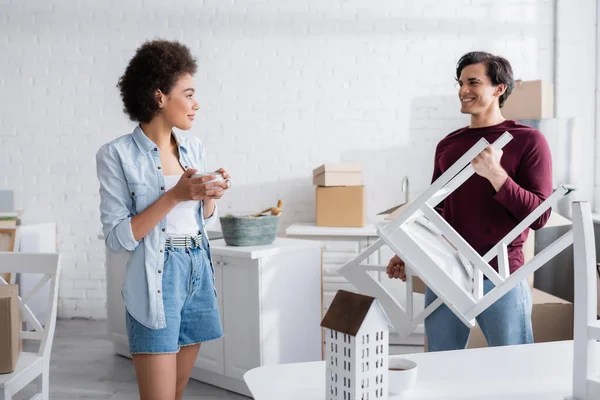 Mujer afroamericana feliz sosteniendo taza de té y mirando novio con silla - foto de stock