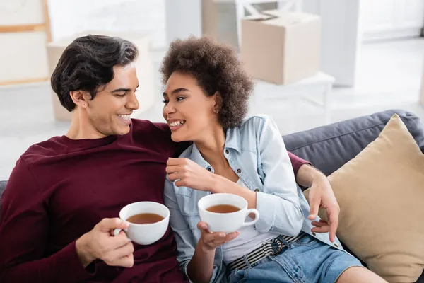 Alegre pareja multiétnica sosteniendo tazas de té mientras descansa en el sofá - foto de stock