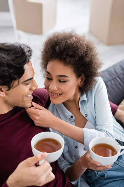 Complacida pareja multiétnica sosteniendo tazas de té mientras descansa en el sofá - foto de stock