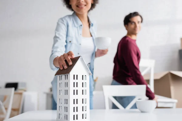 Feliz africana americana mujer celebración taza de té y llegar a casa modelo cerca borrosa novio con silla - foto de stock