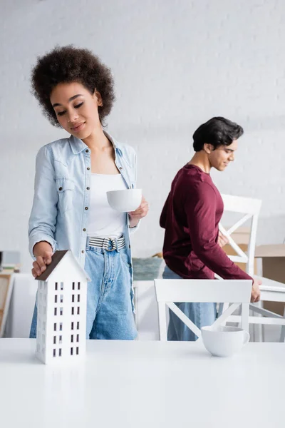 Lächelnde Afroamerikanerin hält Tasse Tee in der Hand und erreicht Hausmodell in der Nähe von verschwommenem Freund mit Stuhl — Stockfoto