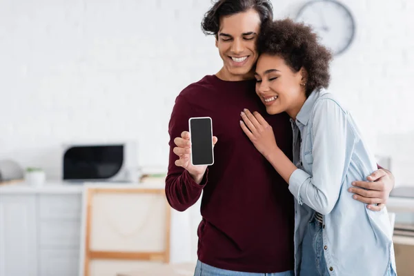 Glücklicher Mann hält Smartphone mit leerem Bildschirm neben lächelnder afrikanisch-amerikanischer Freundin — Stockfoto