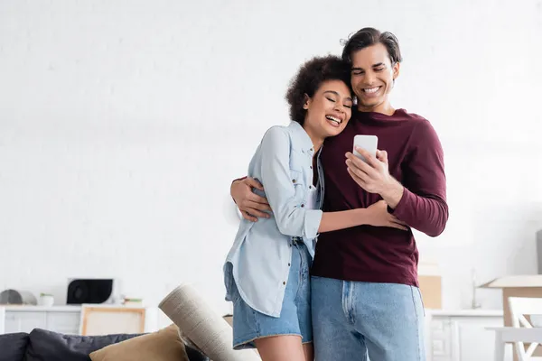 Homem feliz usando smartphone e abraço sorridente afro-americano namorada — Fotografia de Stock