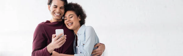 Homme heureux en utilisant smartphone et étreignant souriant petite amie afro-américaine, bannière — Photo de stock