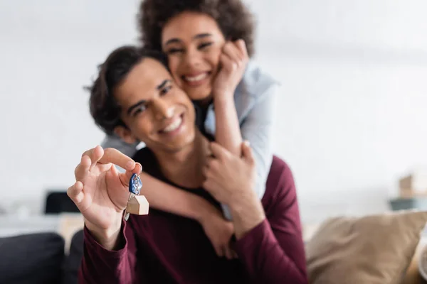 Hombre feliz sosteniendo la llave de un nuevo hogar mientras se abraza con una novia afroamericana sonriente - foto de stock