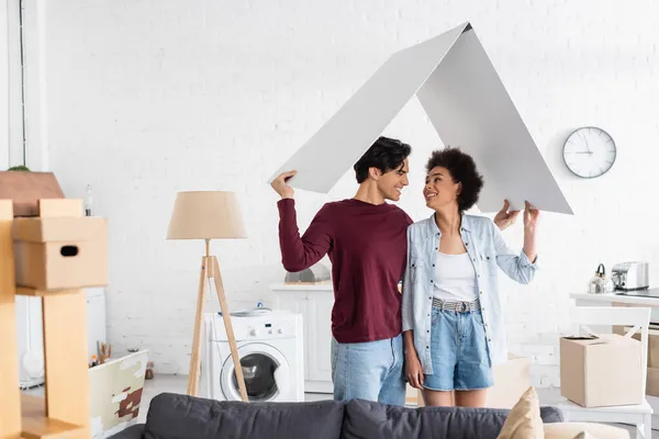 Hombre feliz y sonriente mujer afroamericana sosteniendo techo de cartón en nuevo hogar - foto de stock