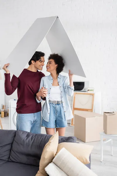 Hombre feliz y sonriente mujer afroamericana sosteniendo techo de cartón en nuevo hogar - foto de stock