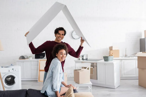 Happy man holding carton roof near smiling african american girlfriend — Stock Photo