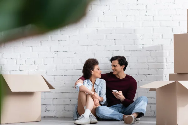 Glückliches multiethnisches Paar, das auf dem Boden sitzt und einander in der Nähe von Kartons ansieht — Stockfoto